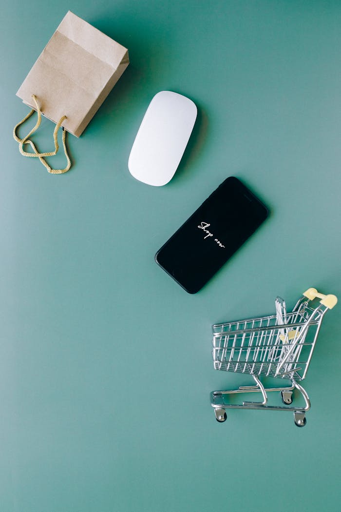 services-04 Top view of a smartphone, mouse, paper bag, and mini shopping cart on a green background, symbolizing e-commerce.
